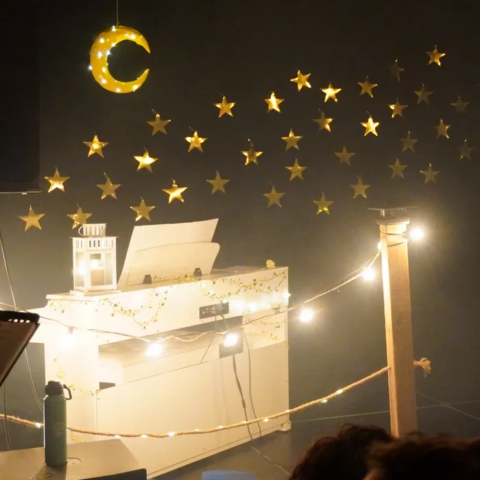 Un piano blanc orné de guirlandes lumineuses, sous une lune et des étoiles dorées.